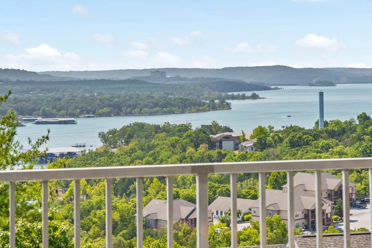 A wide, serene view of Table Rock Lake with a lush green landscape and a hint of residential areas. The horizon blends the calm water and sky, evoking a sense of tranquility and endless outdoor adventure. The setting suggests opportunities for boating, fishing, and peaceful relaxation on a balcony or lakeside.