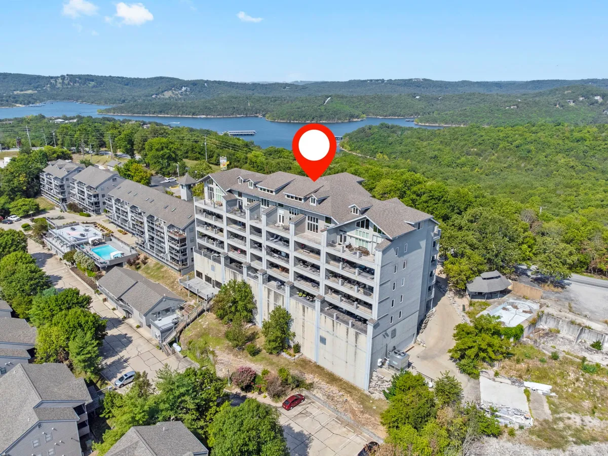 A wide-angle exterior shot featuring a multi-story building with balconies, lush greenery, a view of the pool, and the sparkling lake beyond. The composition brings together the tranquility of the lake, the convenience of the pool, and the comfort of condo living.