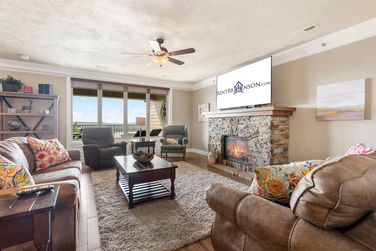 The image showcases a warm, inviting living area featuring a beautiful stone fireplace, expansive windows that flood the space with natural light, plush comfortable seating arranged for conversation or relaxation, a central coffee table, and a television for entertainment. The overall ambiance is cozy and ideal for family gatherings, relaxing evenings, or enjoying the lake views from indoors.
