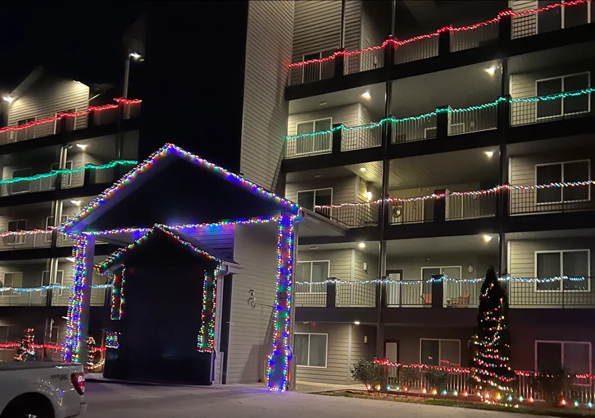 A festive exterior shot showcasing colorful holiday lights, a decorated entrance, and a prominent Christmas tree. The scene radiates cheer and activity, suggesting communal gatherings and holiday celebrations just outside the property.
