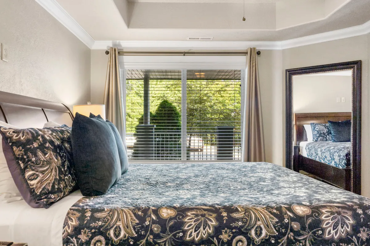 A serene bedroom featuring a king-sized bed with intricate bedding, large windows filling the room with natural light, and a mirror reflecting the tranquil space. The room exudes a sense of restful luxury and comfort.